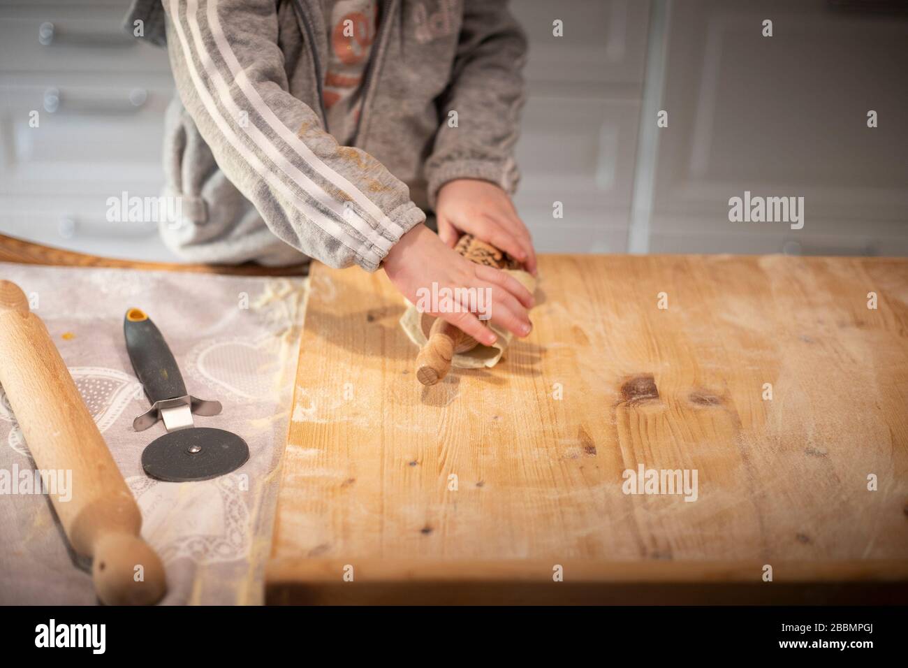 Child girl hands, in white kitchen, flattening pizza dough with a rolling pin on a wooden board. Detail. Lockdown activity idea. Stock Photo