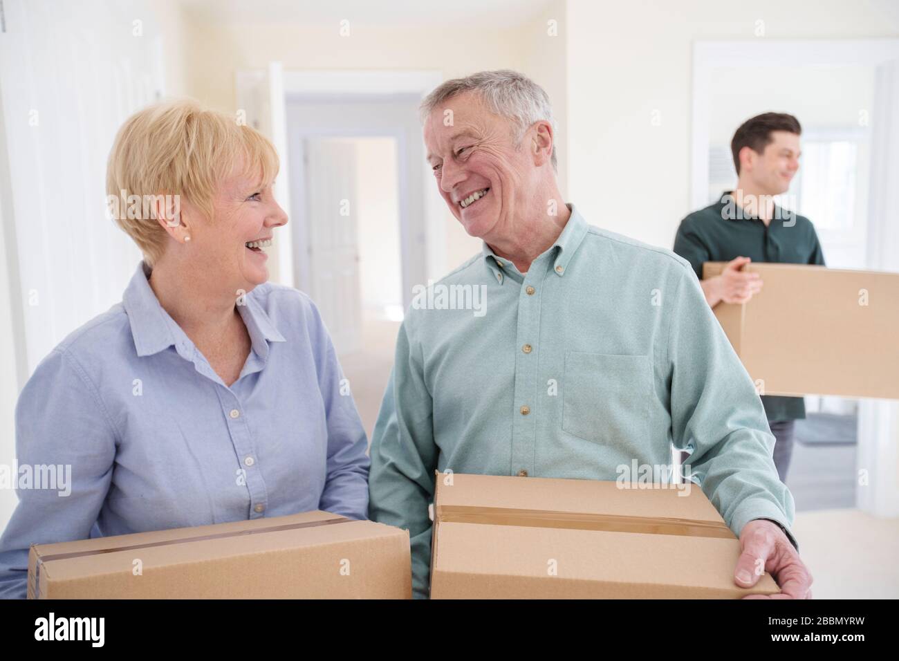 Senior Couple Downsizing In Retirement Carrying Boxes Into New Home On Moving Day With Removal Man Helping Stock Photo