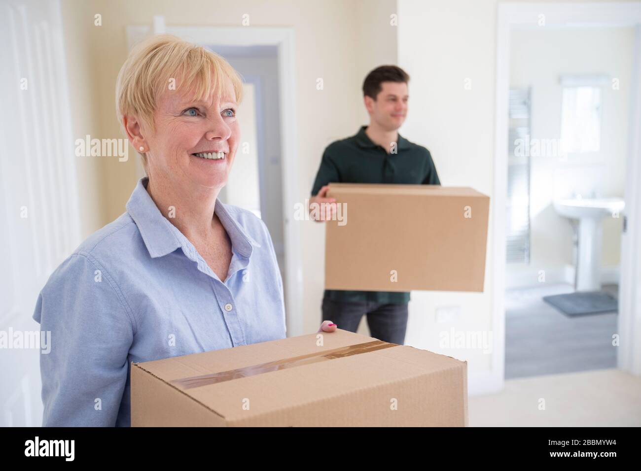 Senior Woman Downsizing In Retirement Carrying Boxes Into New Home On Moving Day With Removal Man Helping Stock Photo