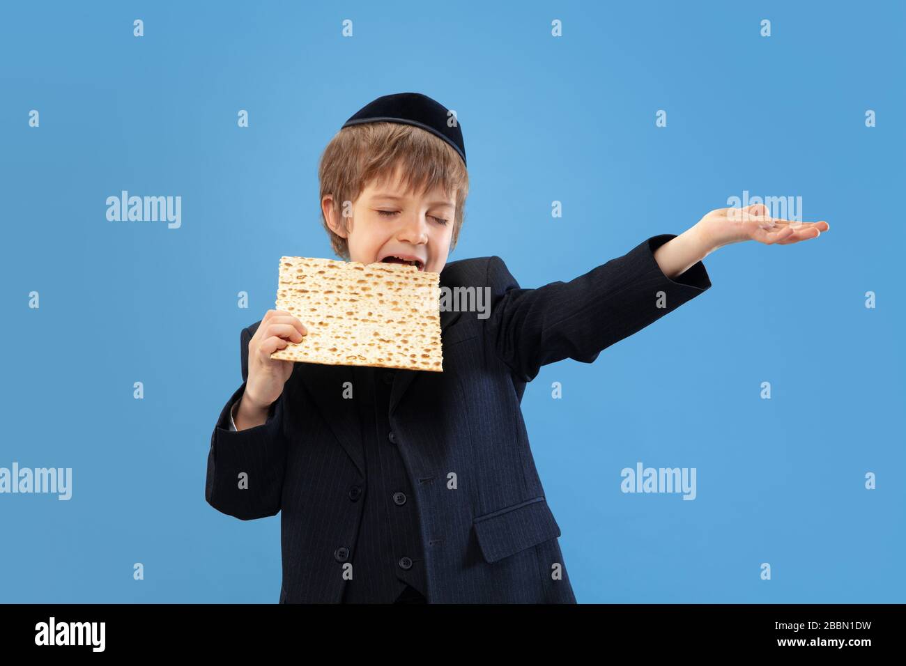 Joyfull eating matzah. Portrait of a young orthodox jewish boy isolated on blue studio background. Purim, business, festival, holiday, childhood, celebration Pesach or Passover, judaism, religion concept. Stock Photo