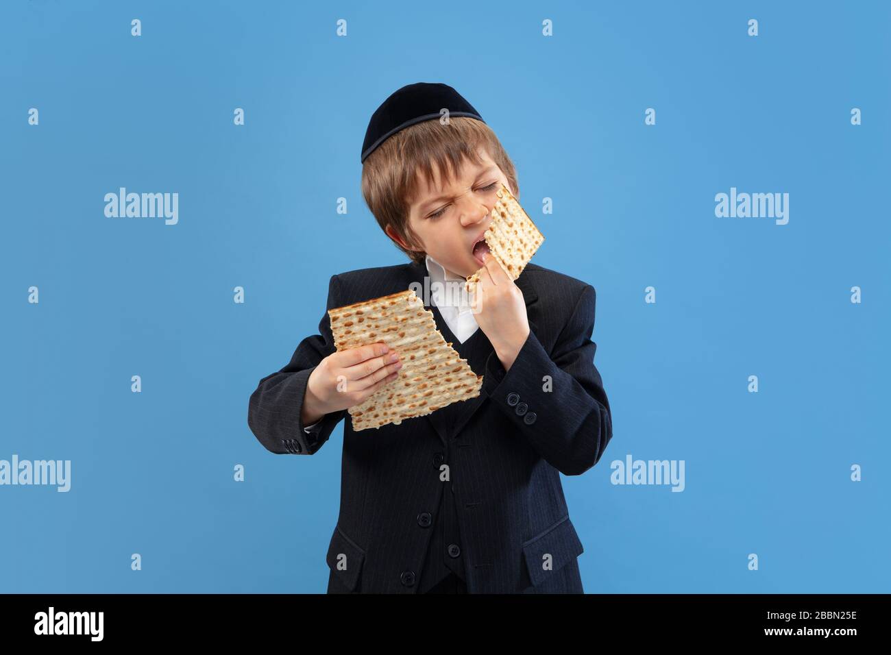 Joyfull eating matzah. Portrait of a young orthodox jewish boy isolated on blue studio background. Purim, business, festival, holiday, childhood, celebration Pesach or Passover, judaism, religion concept. Stock Photo
