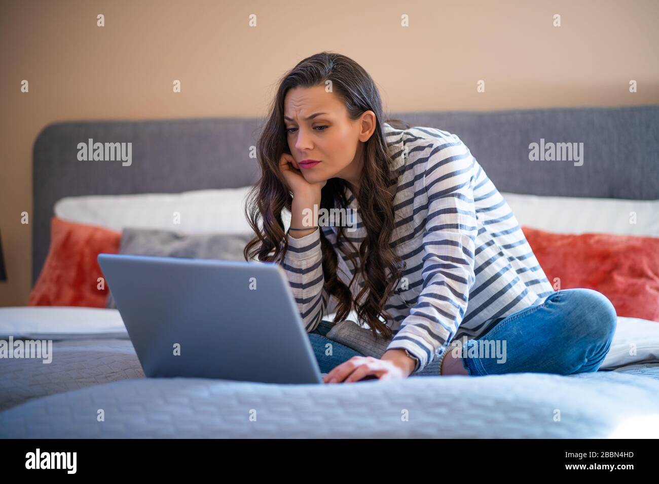Young woman sitting on bed in her home and using laptop. Stock Photo