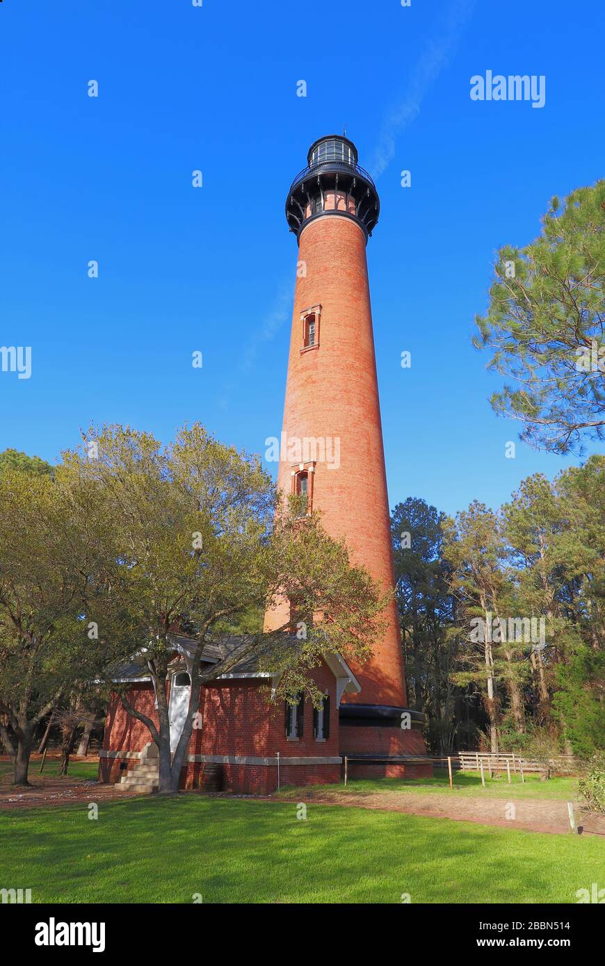 The red brick structure of the Currituck Beach Lighthouse rises overover a small entry house and pine trees at Currituck Heritage Park near Corolla, N Stock Photo