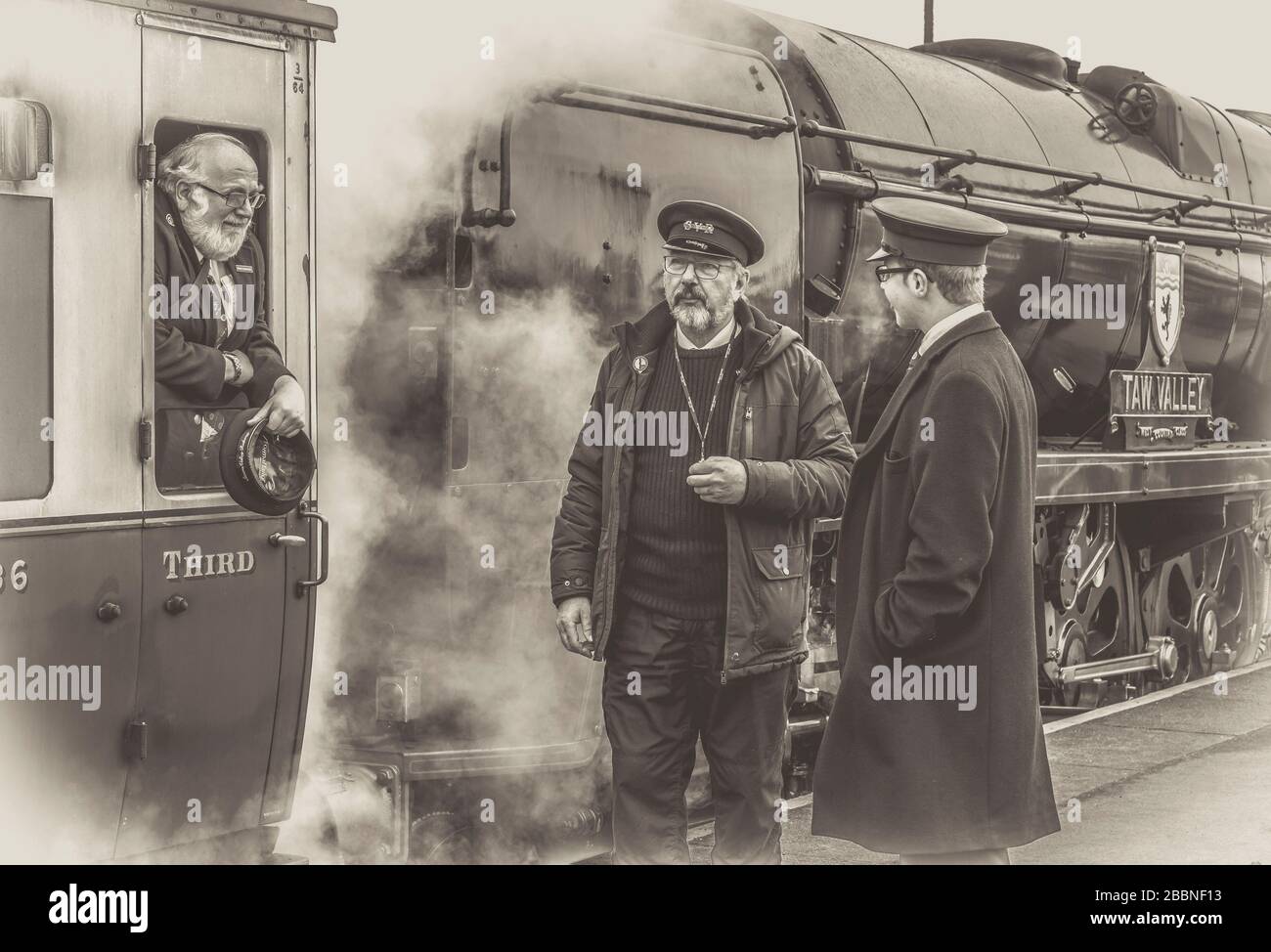 Black & white view railway workers chatting, Kidderminster station platform, Severn Valley heritage railway. Guard leaning out of vintage steam train. Stock Photo