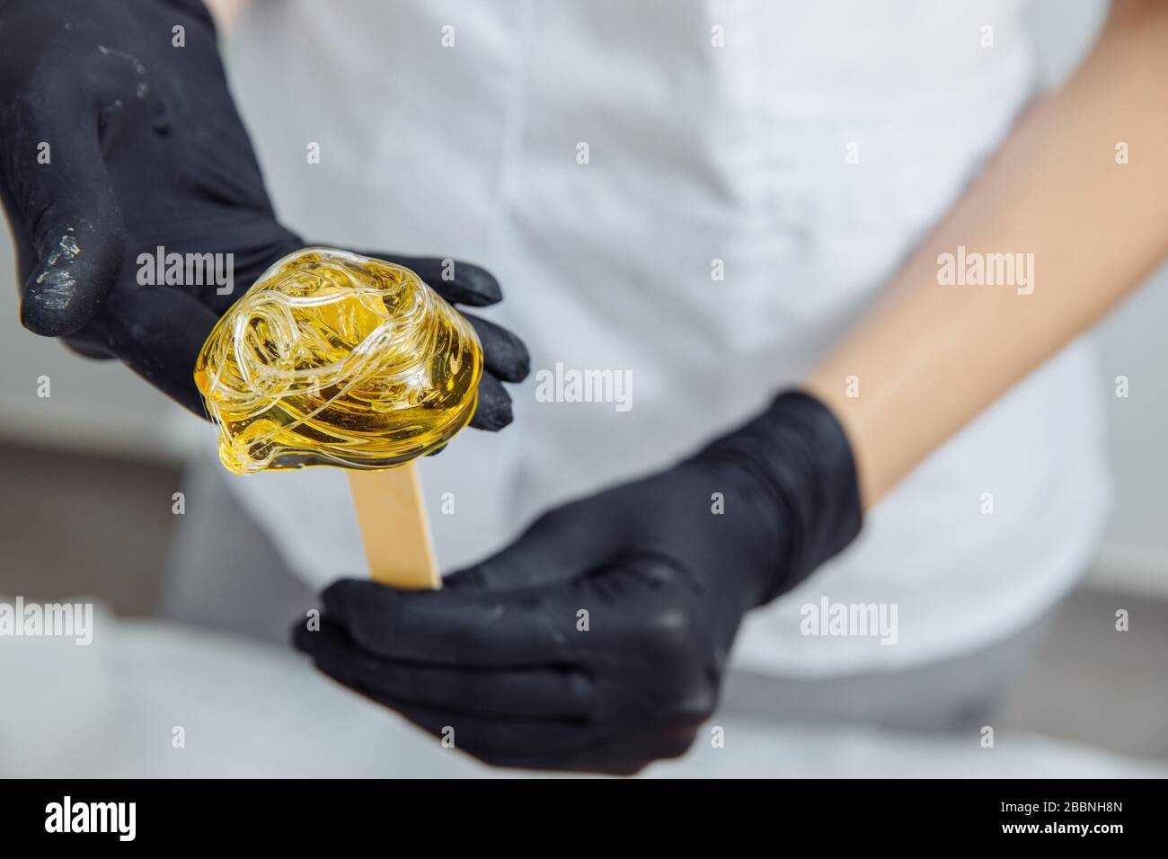 The wax is wound on a wooden spatula. The wax is intended for depilation. The spatula is held by hands in latex gloves.  Stock Photo