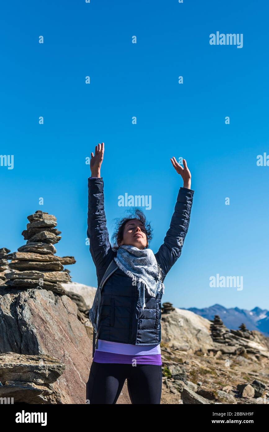 Young white Caucasian woman with her arms up to the sky. Meditating in front of a stone pyramid. Eggishorn terrace, place of energy and power Stock Photo