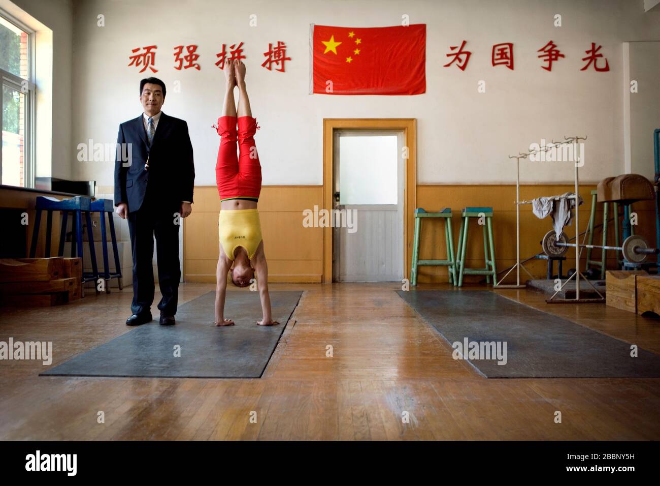Mid-adult man standing beside a teenage girl doing a handstand in a gym. Stock Photo