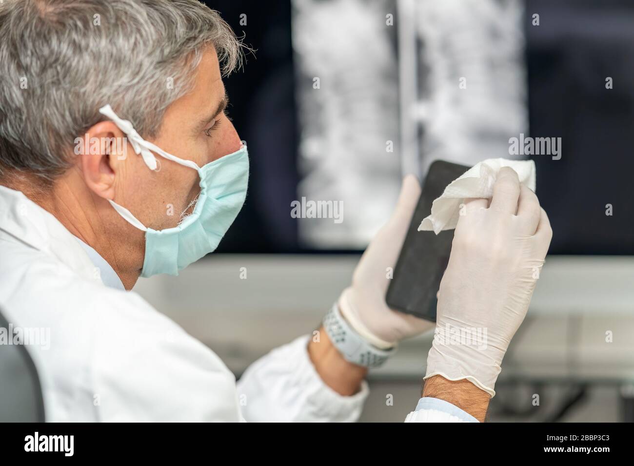 Confident doctor wearing mask and gloves at the medical studio in coronavirus times, cleaning his smartphone. Stock Photo