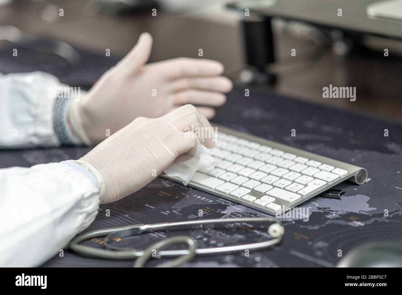Confident doctor wearing mask and gloves at the medical studio in coronavirus times, cleaning his computer keyboard. Stock Photo