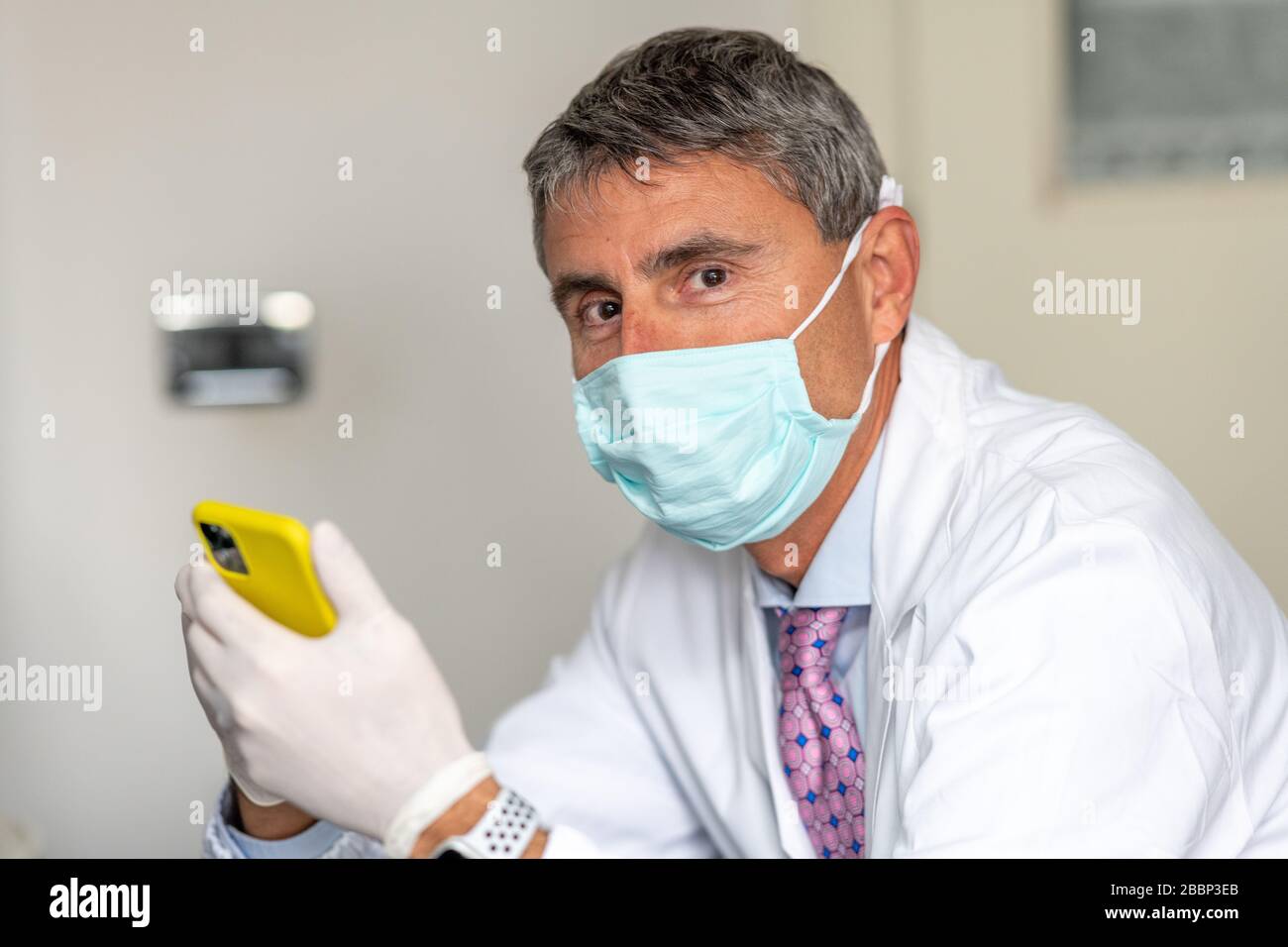 Confident doctor wearing mask and gloves at the medical studio in coronavirus times, cleaning his smartphone. Stock Photo