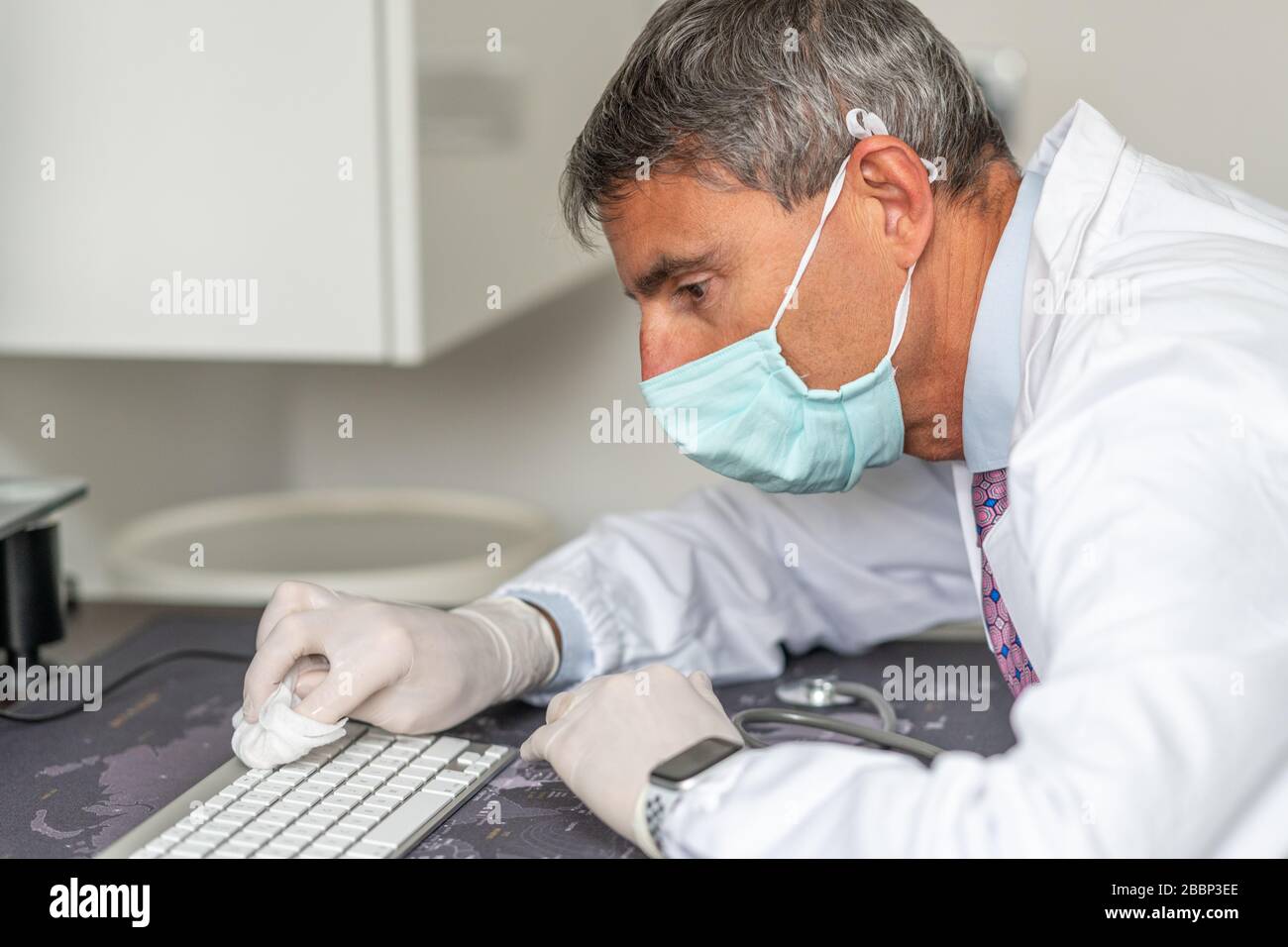 Confident doctor wearing mask and gloves at the medical studio in coronavirus times, cleaning his computer keyboard. Stock Photo