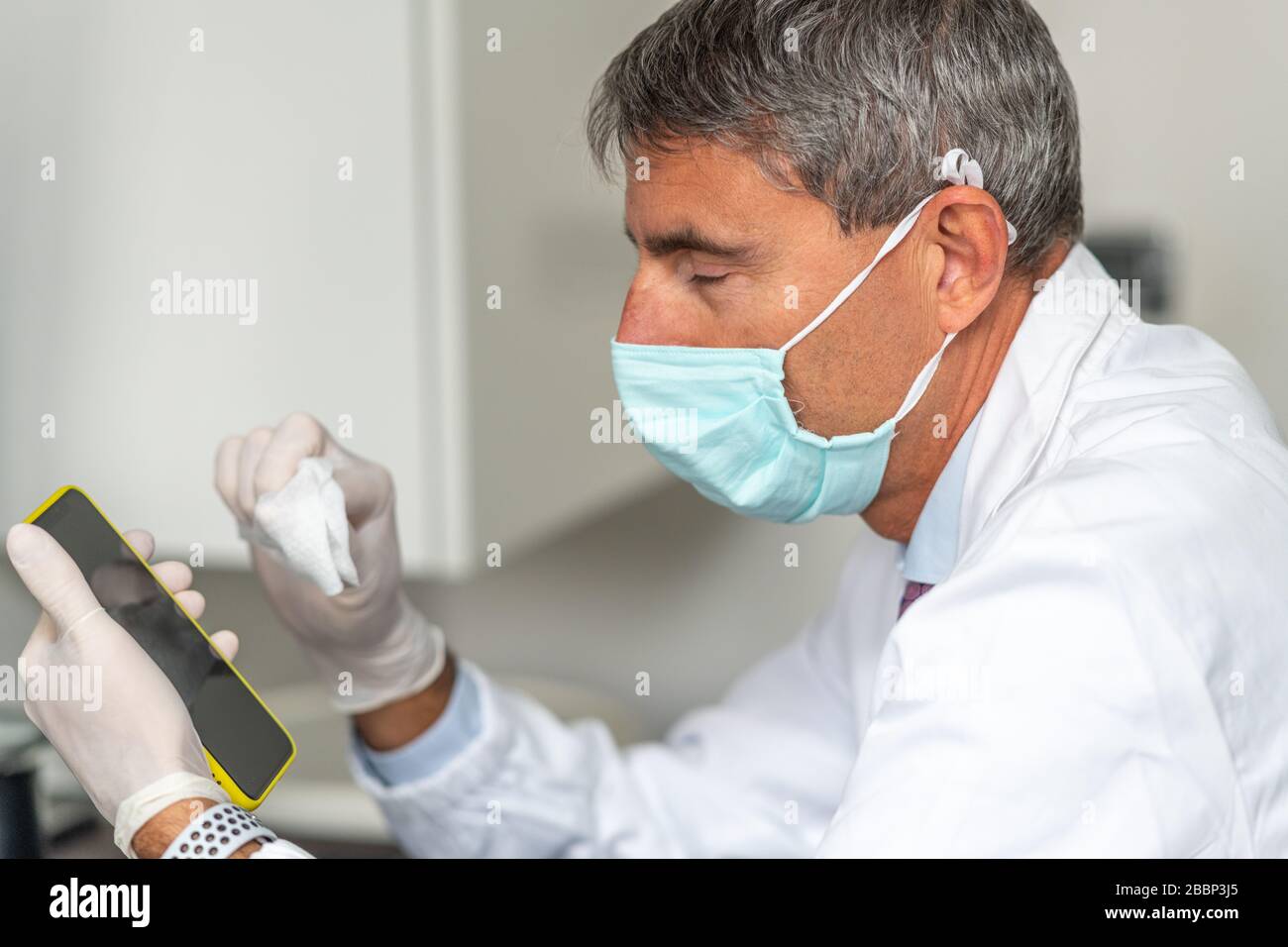 Tired doctor wearing mask and gloves at the medical studio in coronavirus times, cleaning his smartphone. Stock Photo