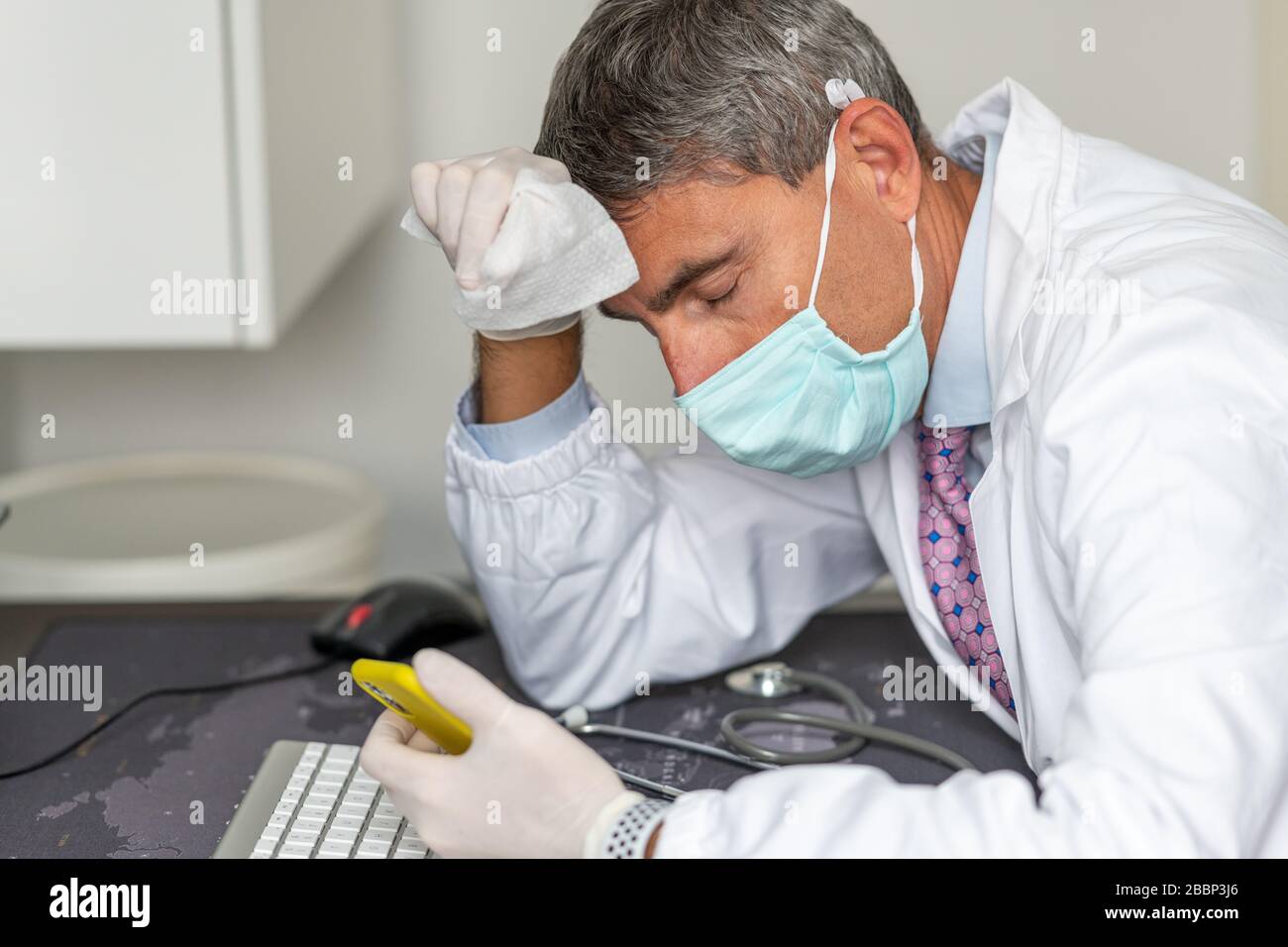 Tired doctor wearing mask and gloves at the hospital in coronavirus times, checking his smartphone. Stock Photo