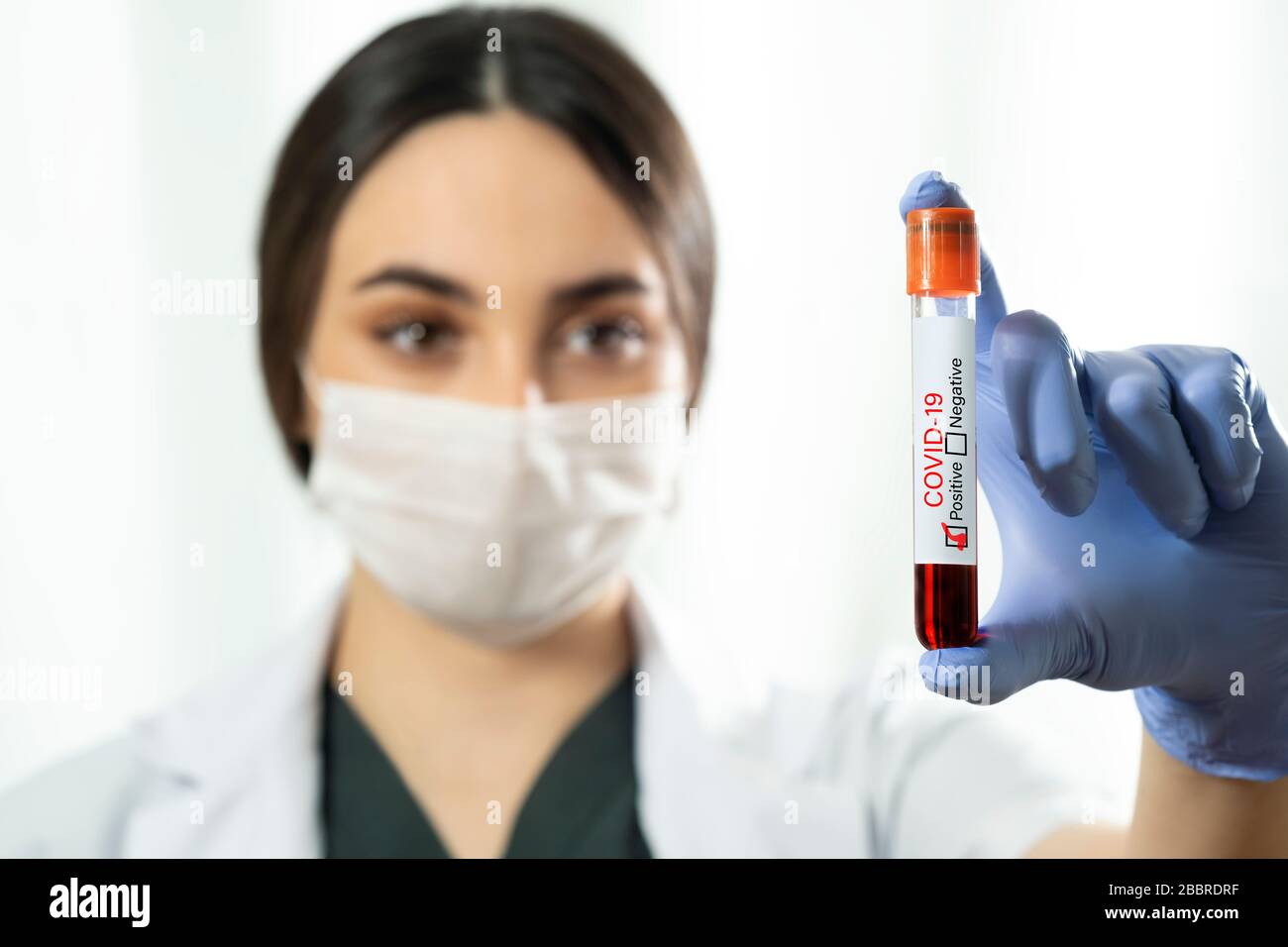Caucasian Woman doctor with surgical mask and blue colored examination gloves holding a blood test tube which is signed as covid-19 positive on a whit Stock Photo