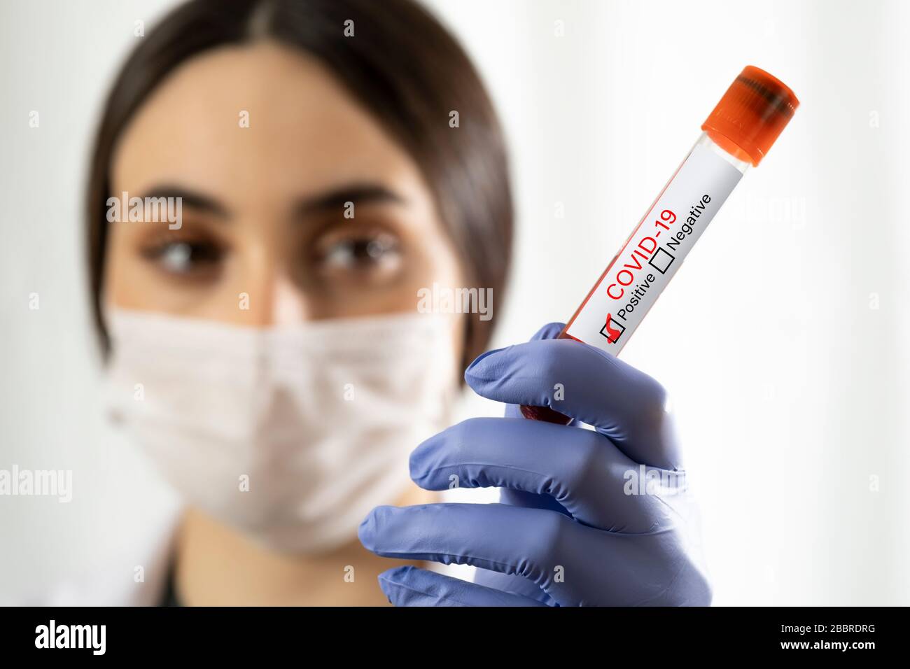 Caucasian Woman doctor with surgical mask and blue colored examination gloves holding a blood test tube which is signed as covid-19 positive on a whit Stock Photo