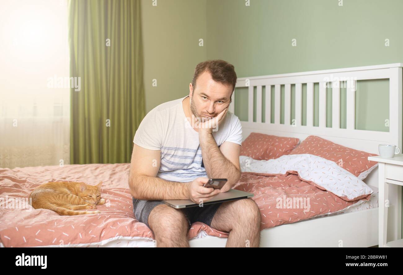 Young man working from home in bedroom, feelling bored and tired holding a phone and laptop in his arms. An orange cat is standing next to him Stock Photo