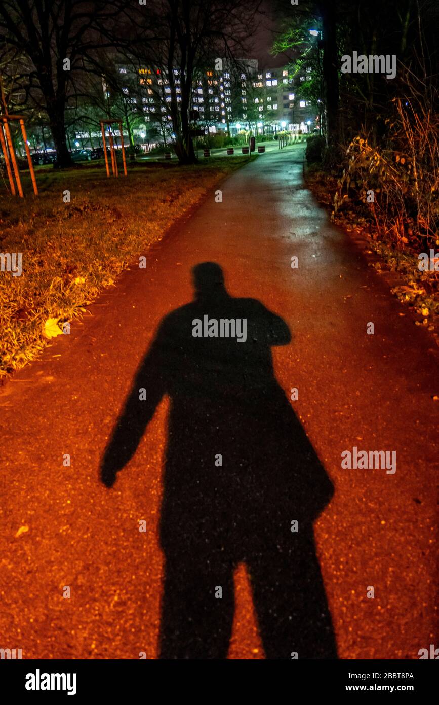 Pedestrian path through a park, in the evening, dark only little lighting by street lamps, shadow of a man, Stock Photo