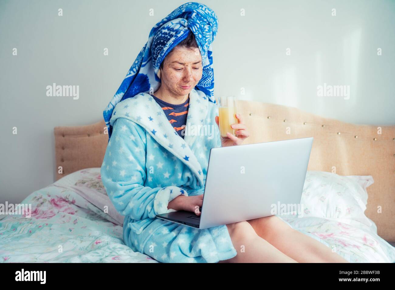 Portrait of a woman in a bathrobe and with a towel on her head, applying a scrubbing mask on her face while using a laptop, sitting on the bed. Lifest Stock Photo