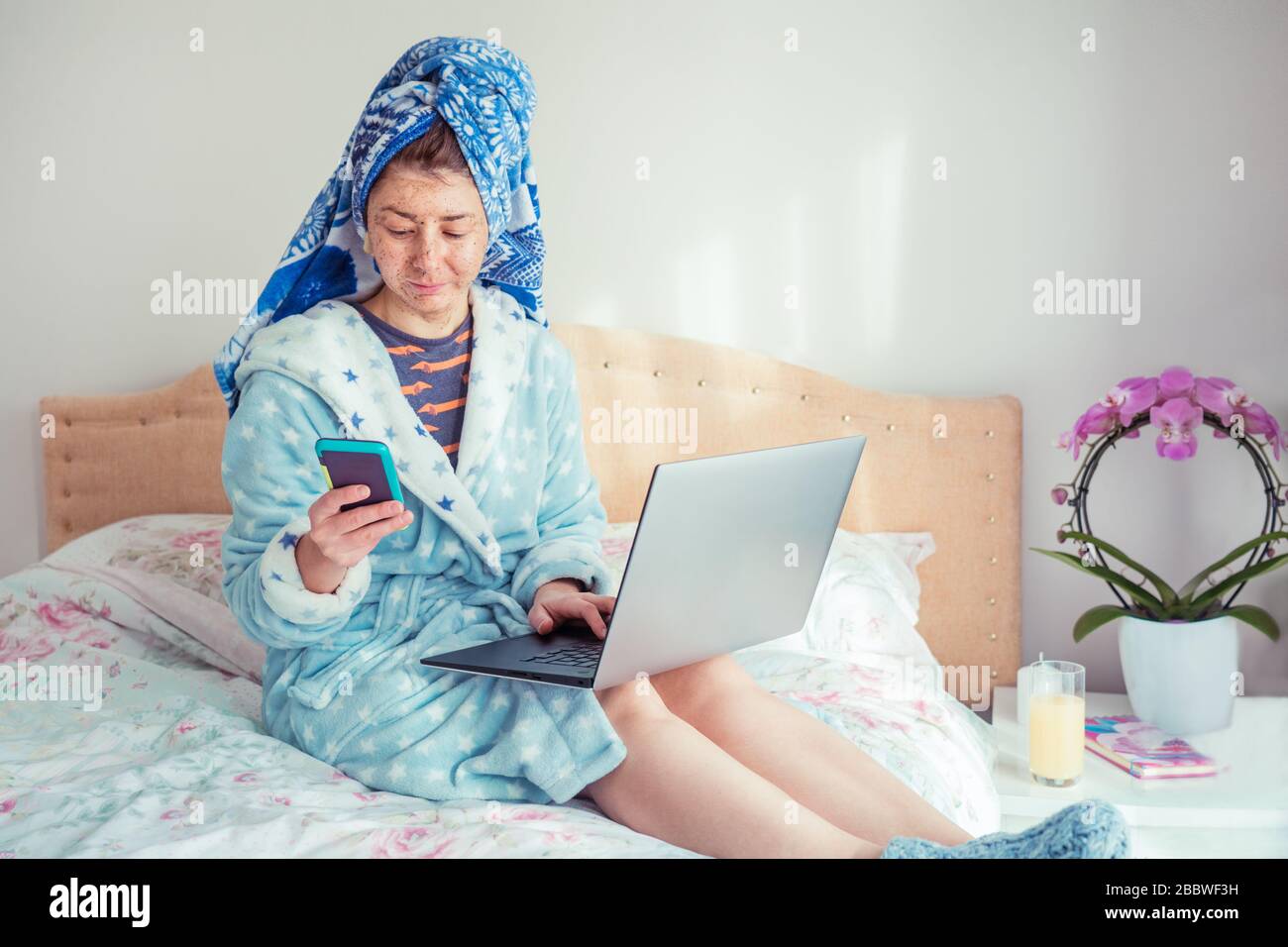 Portrait of a woman in a bathrobe and with a towel on her head, applying a scrubbing mask on her face while using smart phone and laptop, sitting on t Stock Photo