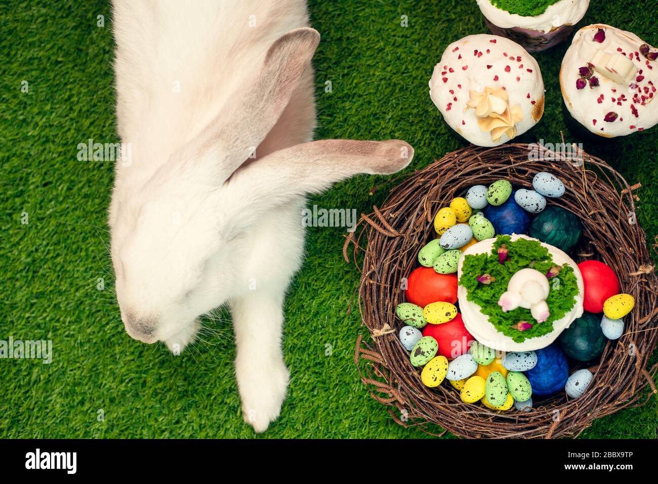 A beautiful fluffy rabbit lies on the green grass next to painted eggs and Easter cakes. Easter holiday concept. Holiday card. Stock Photo