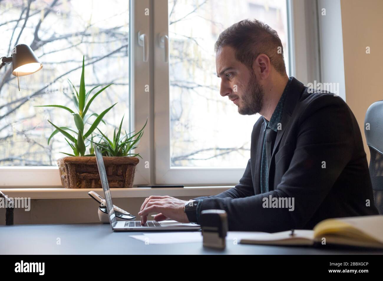 businessman working on laptop in office taking notes by window black table with notebook papers phone Stock Photo