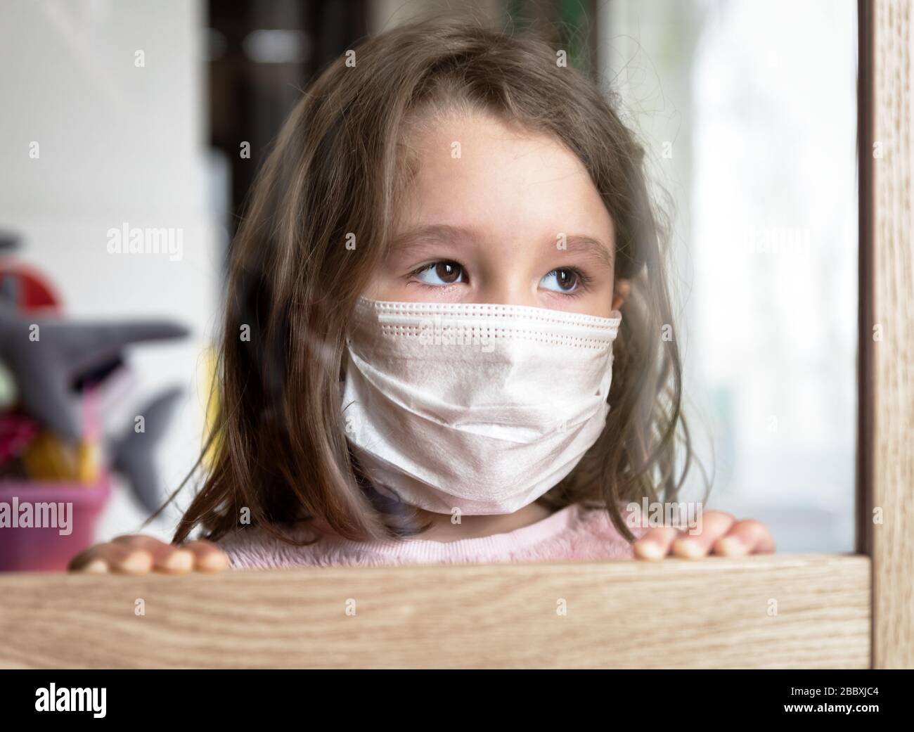COVID-19 coronavirus concept, little girl in face mask looking through window at home or clinic. Portrait of sad kid during quarantine due to coronavi Stock Photo