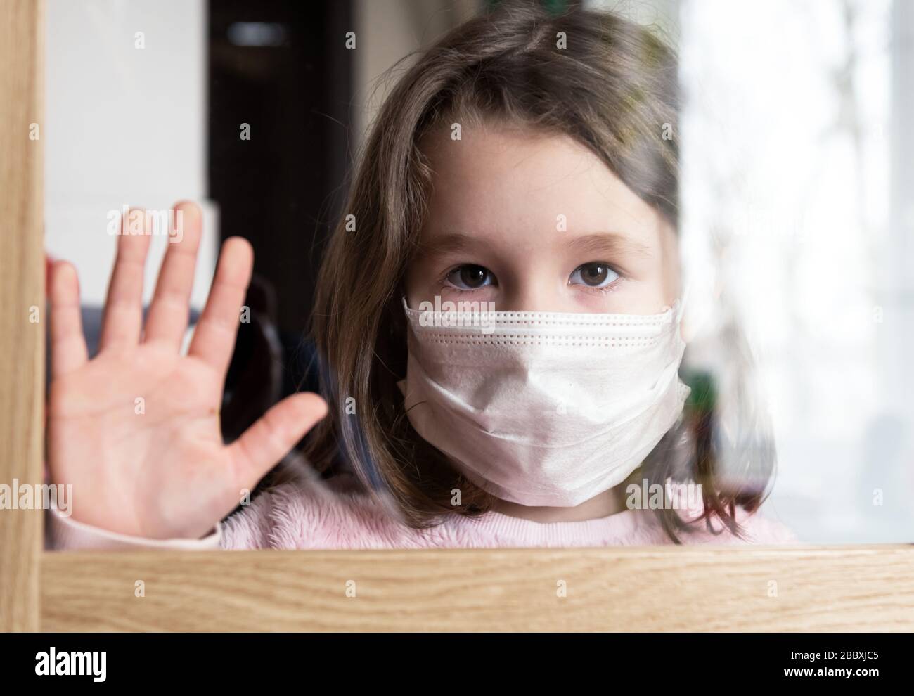 COVID-19 coronavirus concept, little girl in face mask looking through window at home or clinic. Portrait of sad kid during quarantine due to coronavi Stock Photo