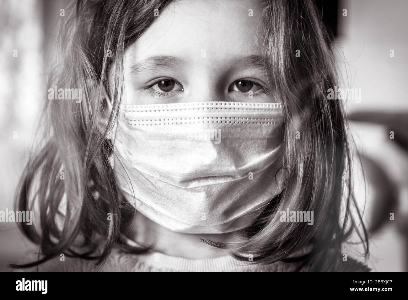 COVID-19 coronavirus concept, little girl in face medical mask looking through window at home or clinic. Portrait of kid during quarantine due to coro Stock Photo