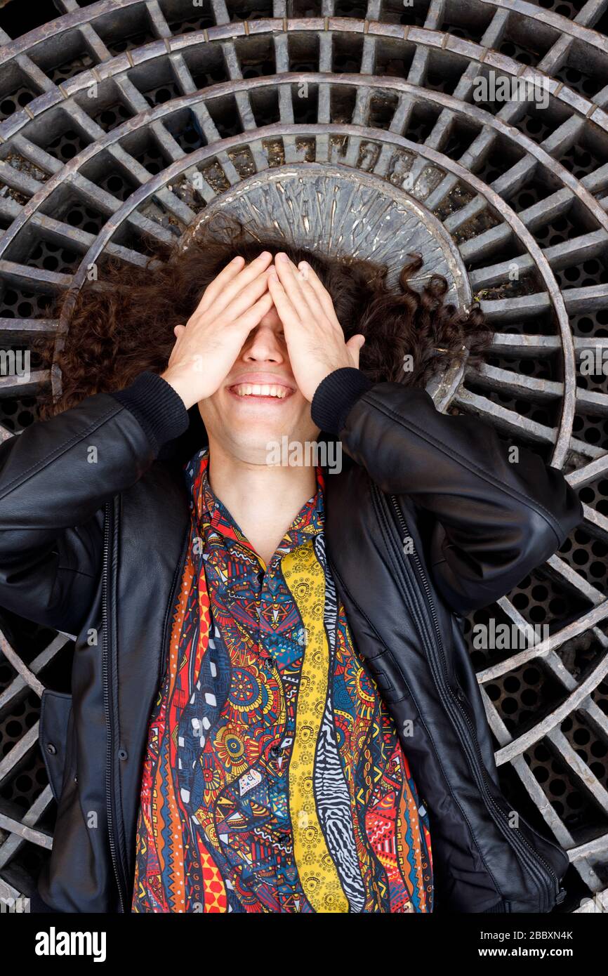 Smiling boy with long hair lying on the ground covering his eyes with his hands Stock Photo