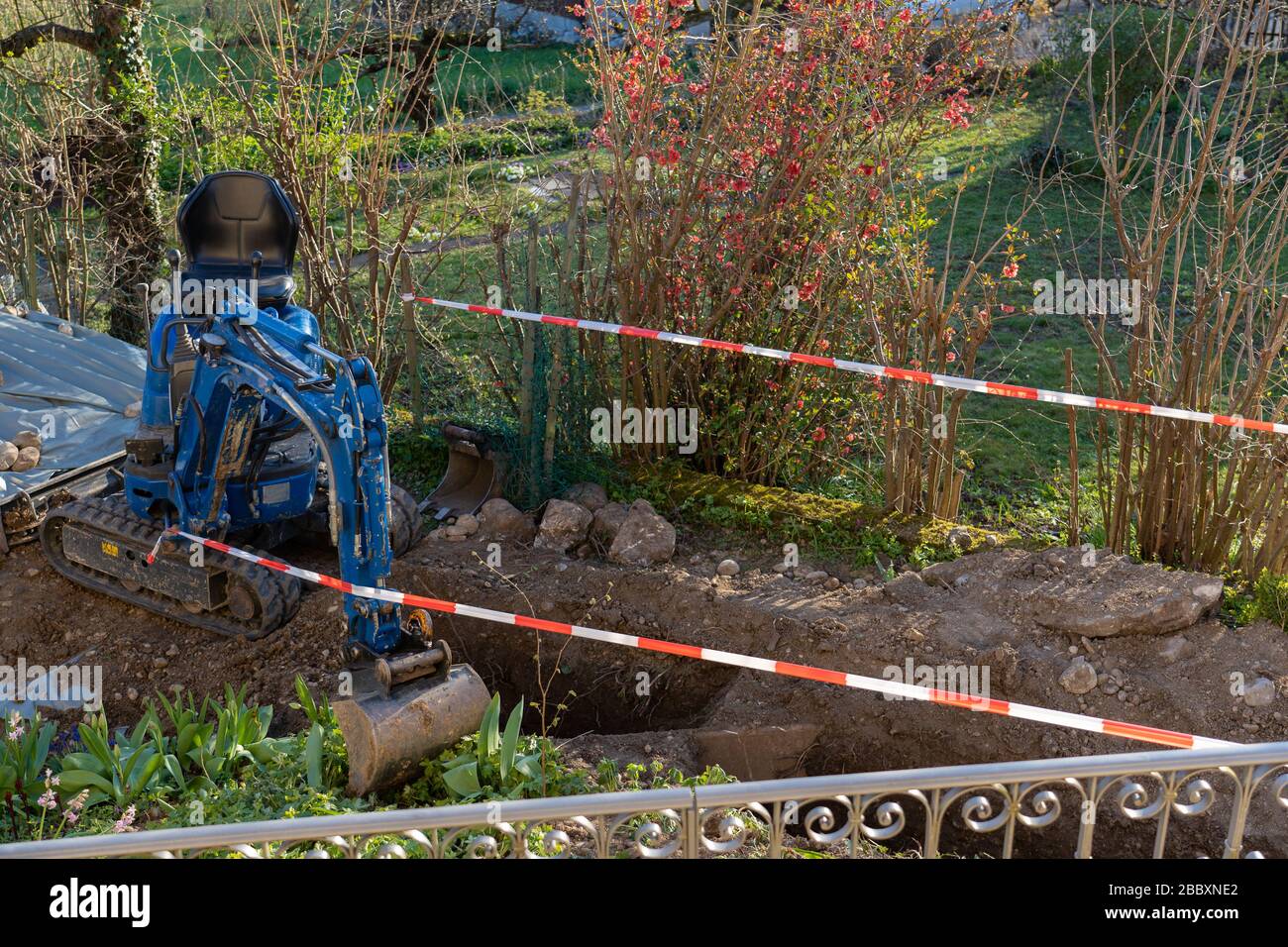 Blue excavator between temporary tape barrier in back yard. Red and white barricade for construction site of sewerage shaft. Stock Photo