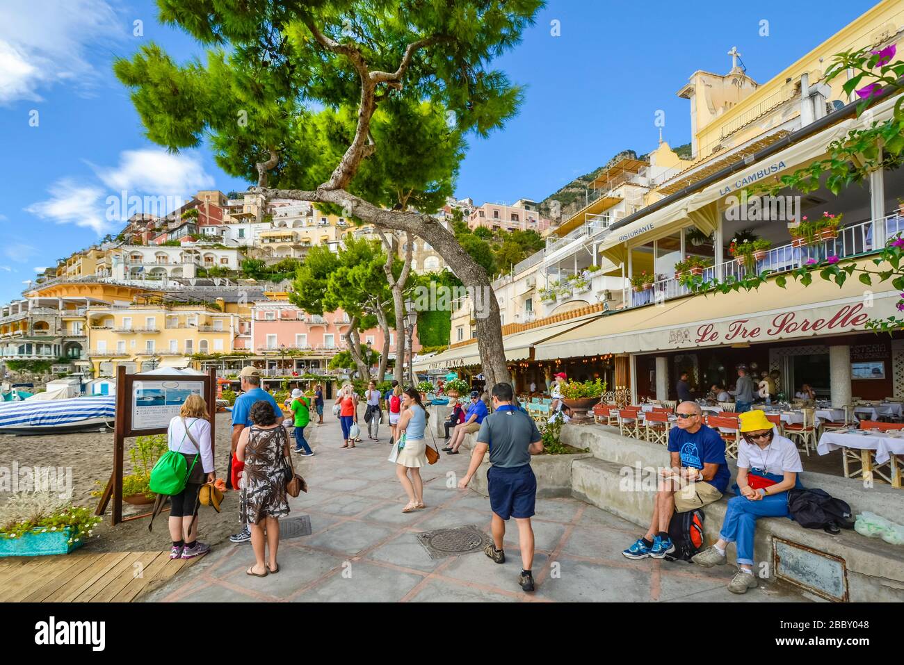 Tourists walk along the boardwalk in front of the sandy beach, cafes and shops at the coastal town of Positano Italy on the  Amalfi Coast. Stock Photo