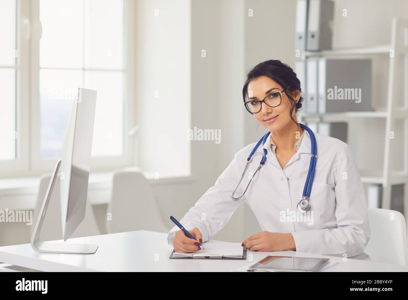 Confident woman doctor pediatrician writes in a clipboard sitting at a table in a white office of the hospital Stock Photo