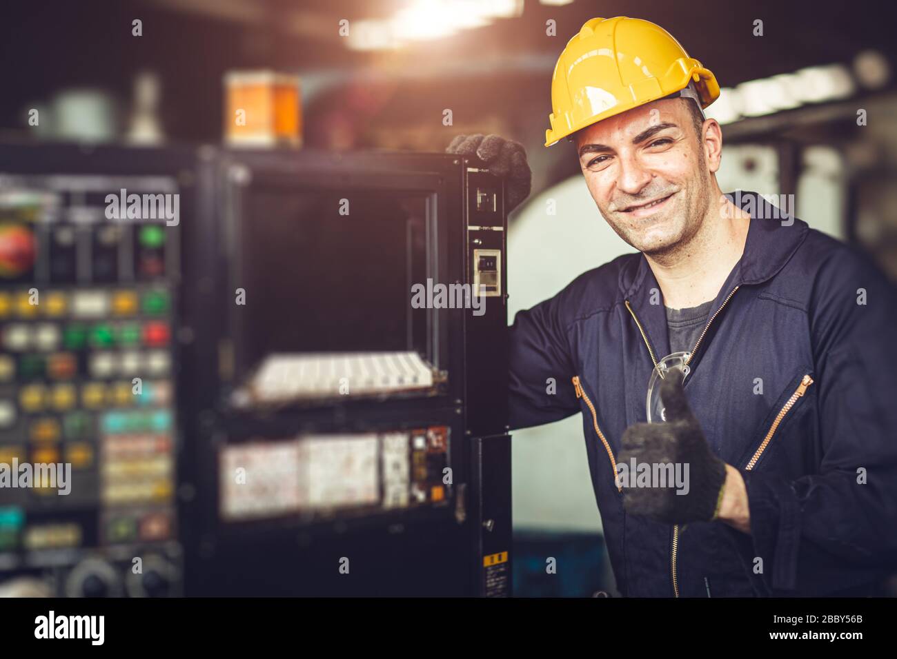 Happy worker, portrait handsome labor thumb signal with safety suit at machine control panel in factory. Stock Photo