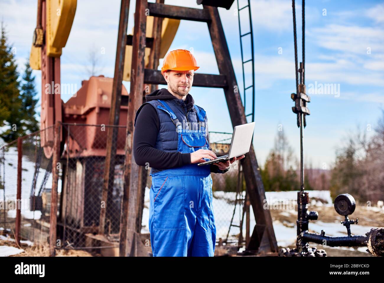 Portrait of petrolium engineer, wearing blue overalls and orange helmet, with a laptop, standing with his back to an oil rig, checking oil pumping unit, making notes in his computer Stock Photo