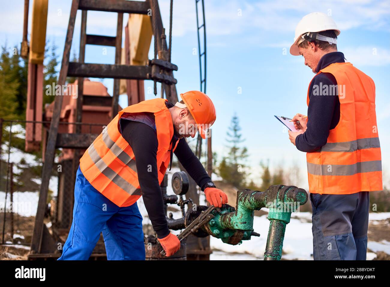 Two oil workers wearing overalls, orange vests, and helmets, working on oil field next to oil pump jack, one is wrenching the pipe, the other making notes. Concept of petroleum industry, collaboration Stock Photo