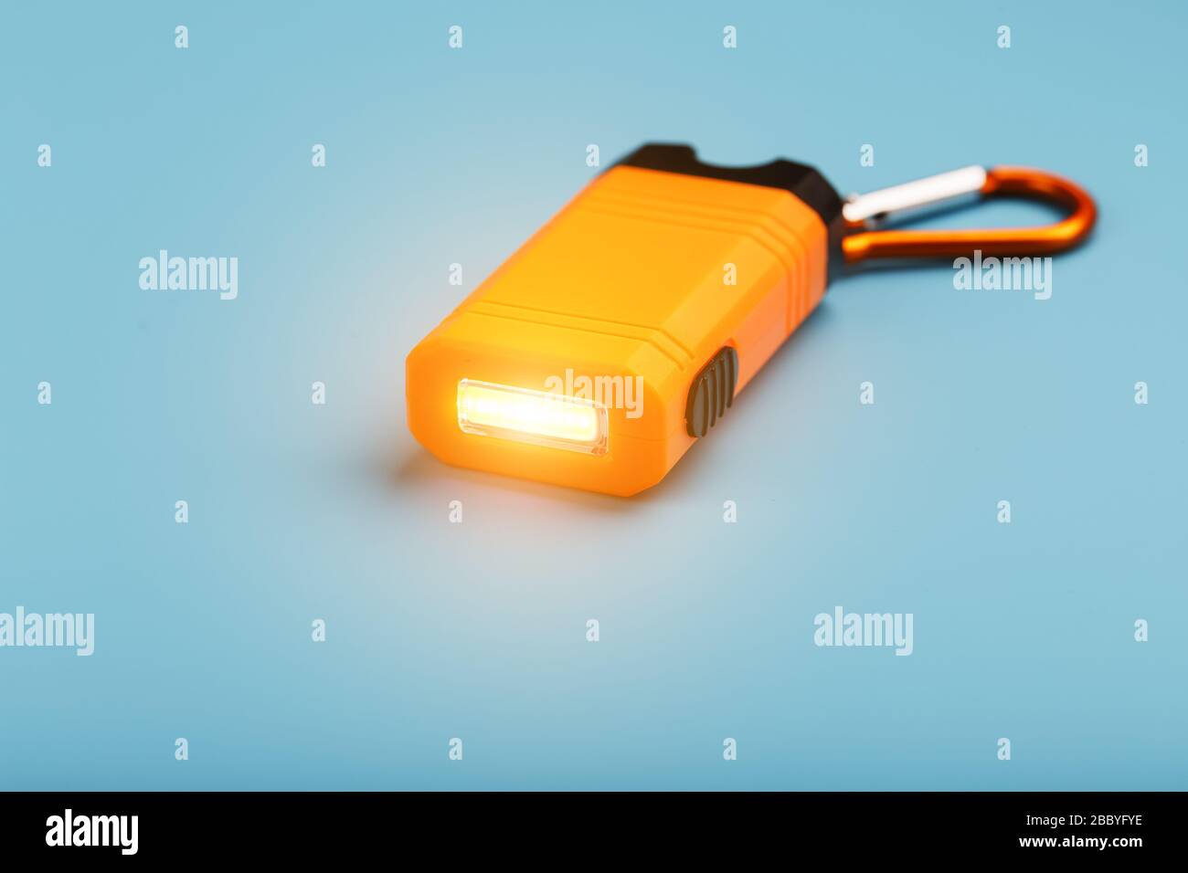 An orange led flashlight with a carabiner glows on a blue background. Stock Photo