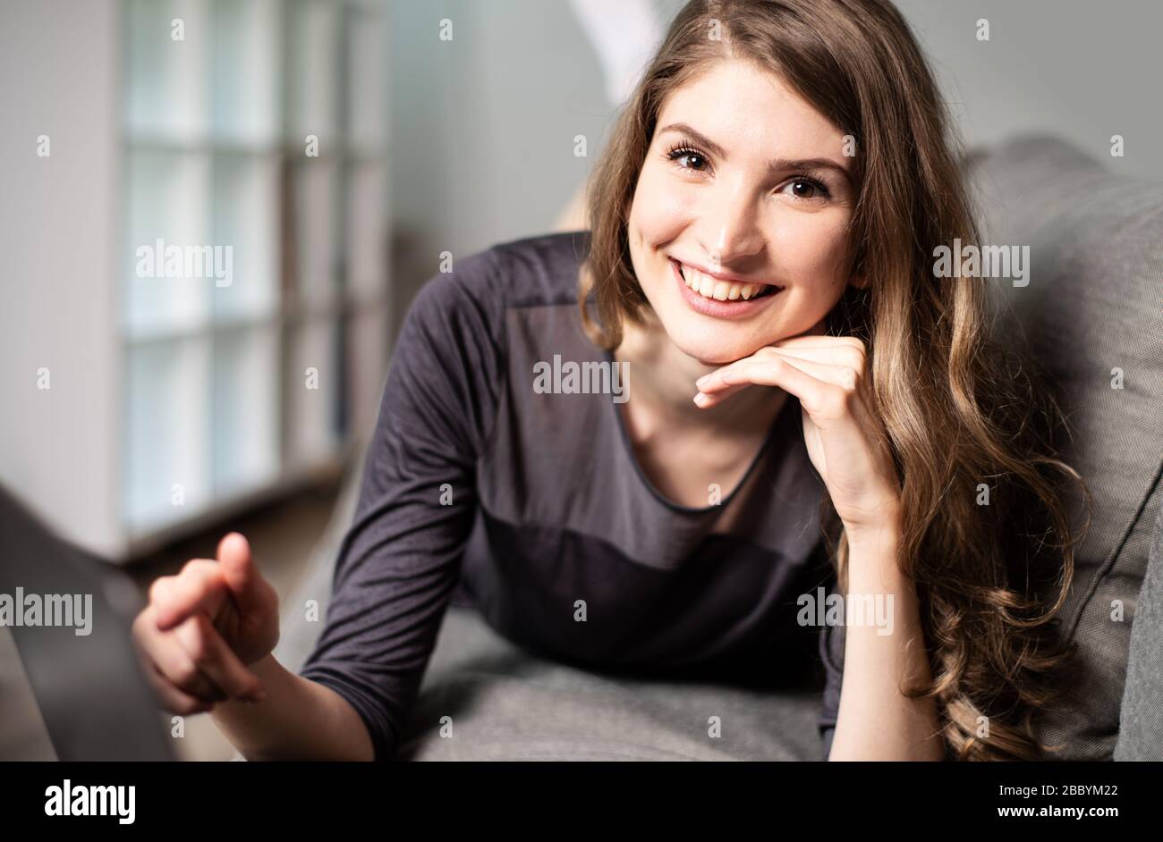 Pretty young woman lying on the couch with a laptop and smiling ...