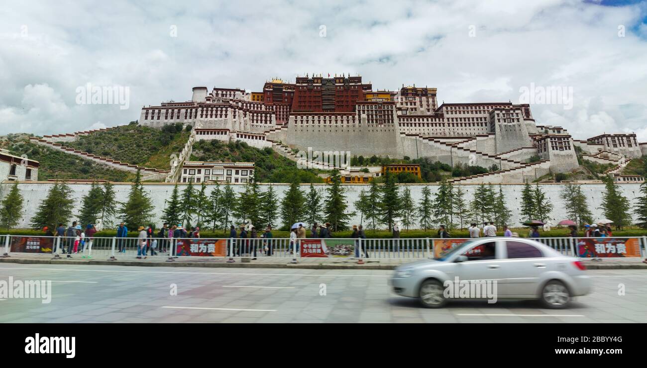 Front view (panorama) of Potala Palace. A silver colored car is passing by from the right to the left. Pedestrians are walking in front of the palace. Stock Photo