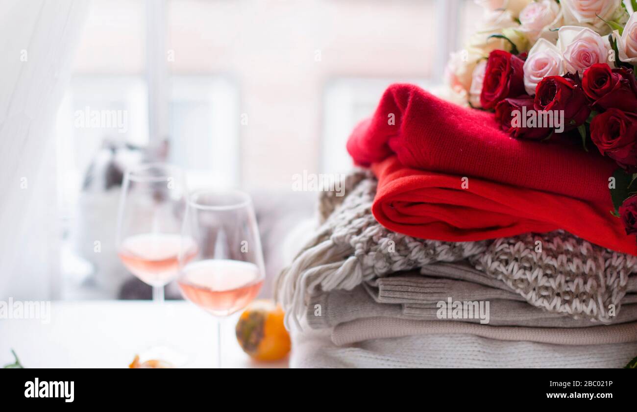 A stack of knitted sweaters and a bouquet of red and white roses near the window. Homeliness, warm clothes,a beautiful cat and two glasses of wine.The concept of gifts and surprises.Banner.Soft focus  Stock Photo