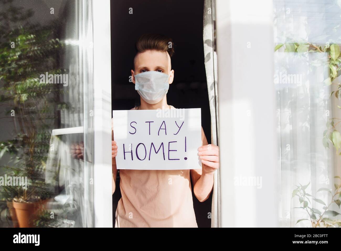 A young man in a protective mask stands at home outside the window and shows a table with the words Stay at home. A call for self-isolation due to a pandemic and the risk of coronavirus infection. Stock Photo