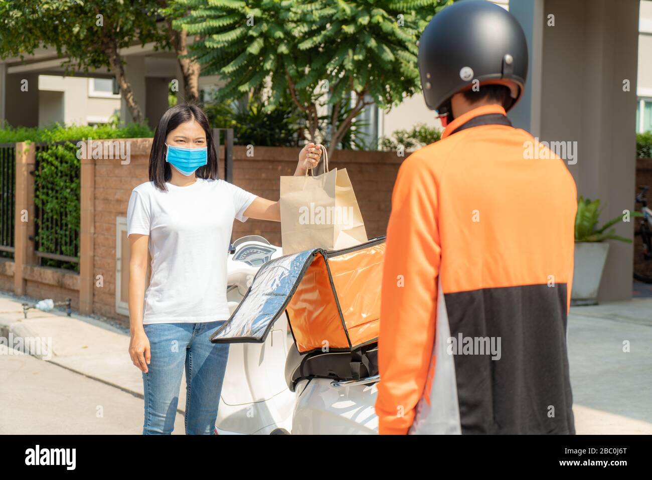 Asian woman pick up delivery food bag from box for contactless or contact free from delivery rider with bicycle in front house for social distancing f Stock Photo