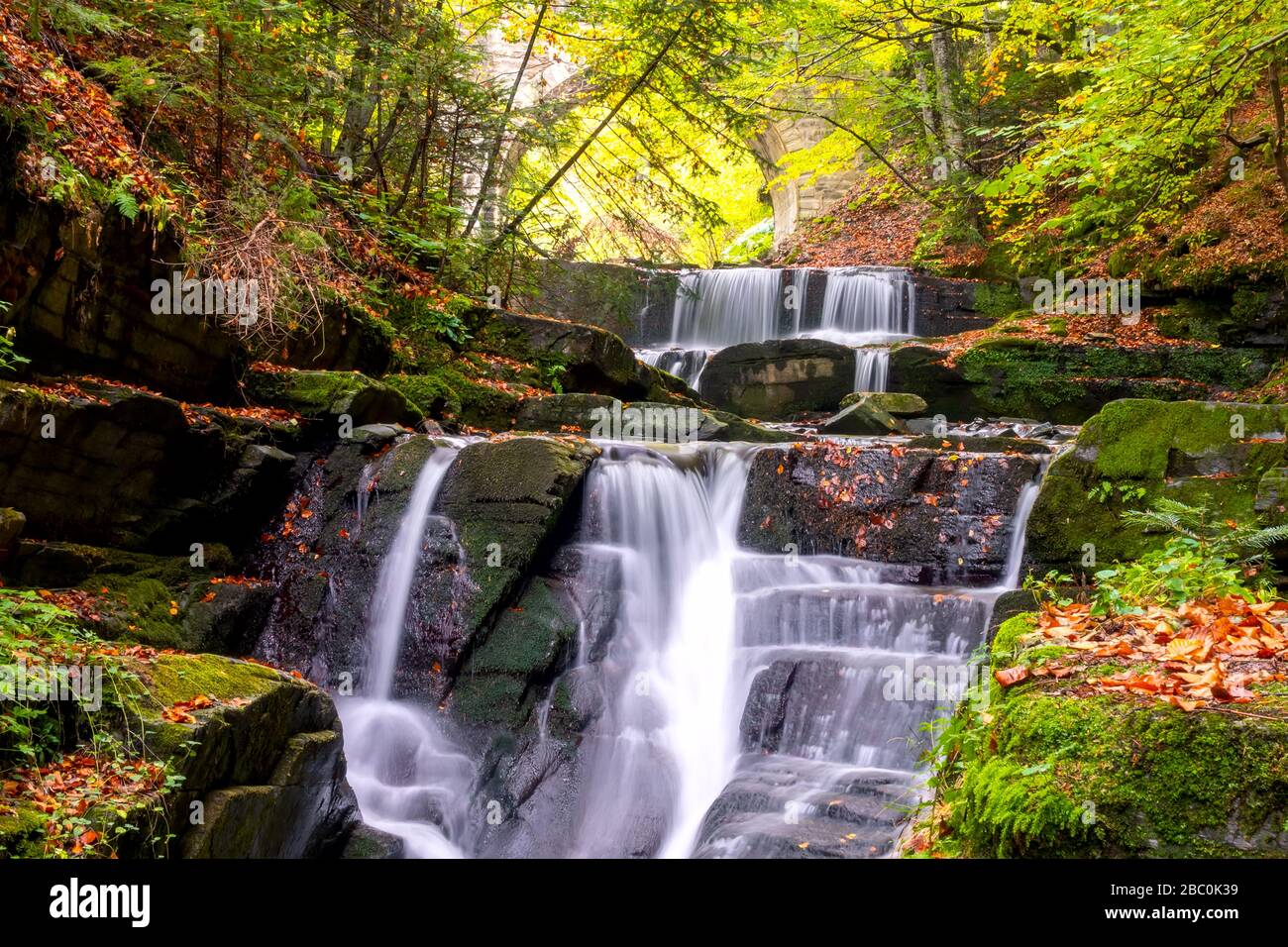 Sunny day in the summer forest. Small river and several natural waterfalls. Arch of an old stone bridge Stock Photo