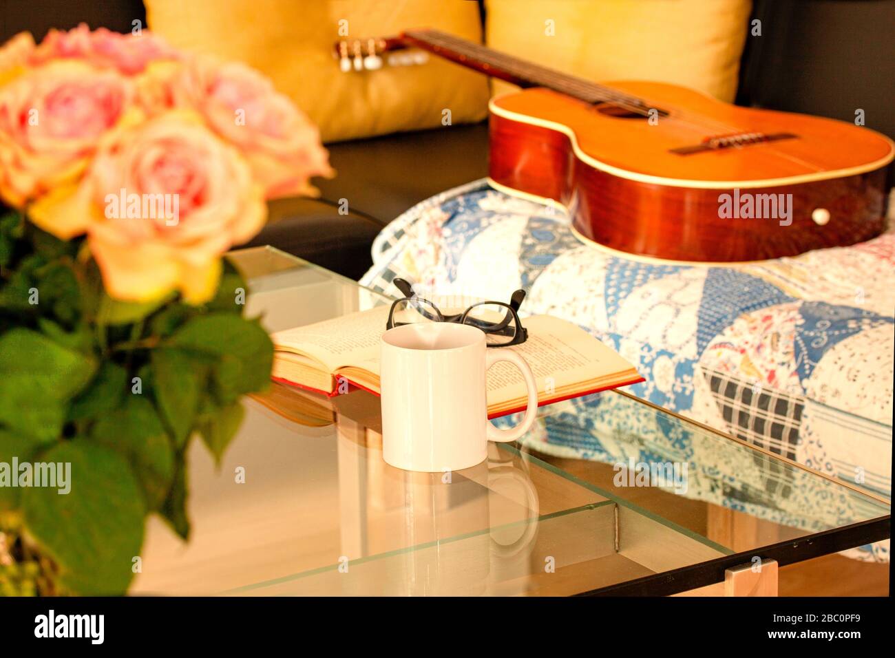 Book, glasses, roses and a mug with tea on modern glass table. An acoustic Spanish guitar is lying on an old rug on a black leather sofa. Living room Stock Photo
