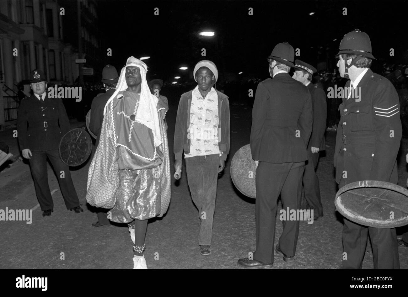 Notting Hill Carnival 1980s. Police use dustbin lids as shield as trouble, begins. Two revellers walk through group of police. 1981  HOMER SYKES Stock Photo