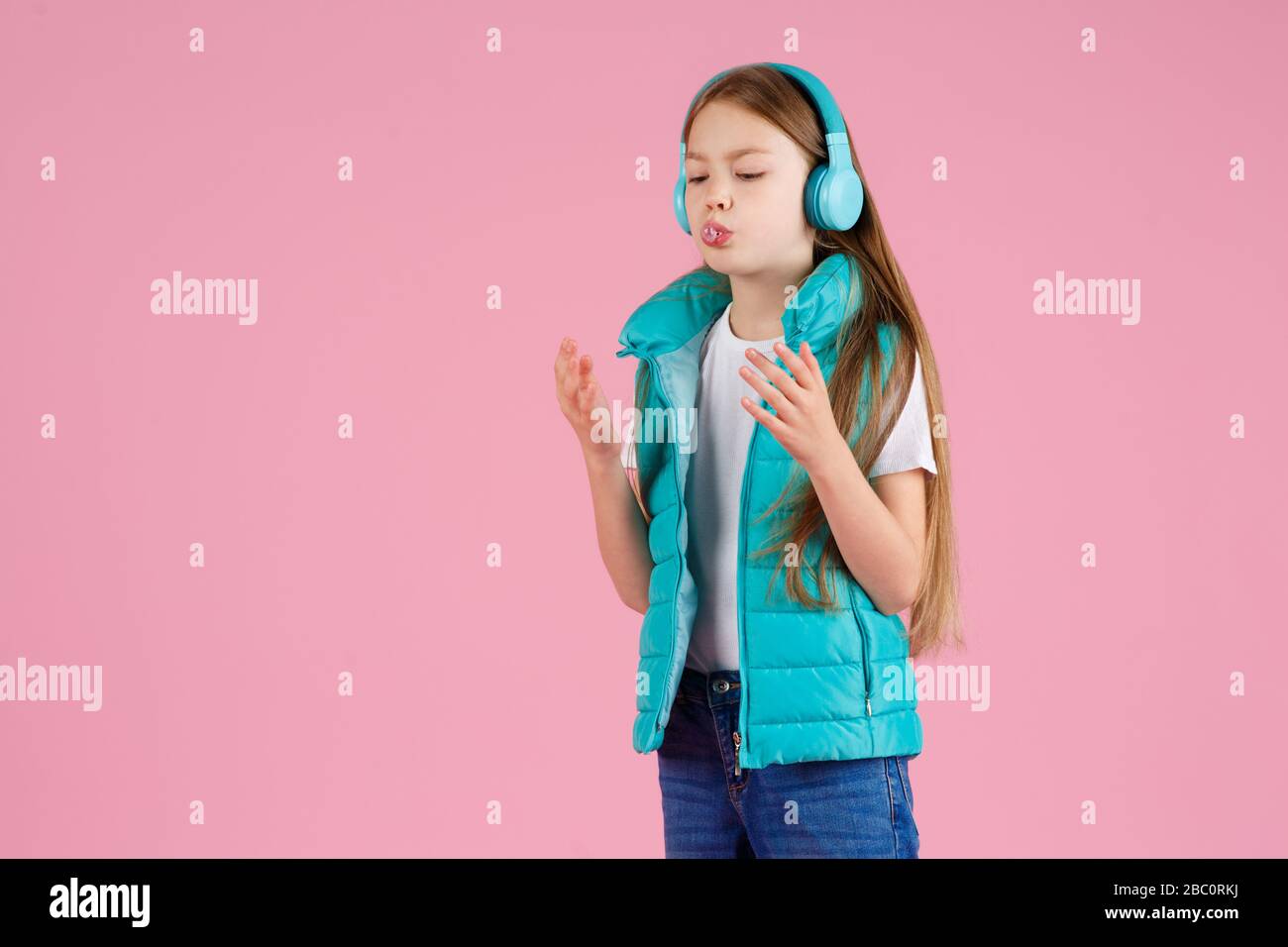 A little girl with headphones explodes pink chewing gum on a pink background. Stock Photo