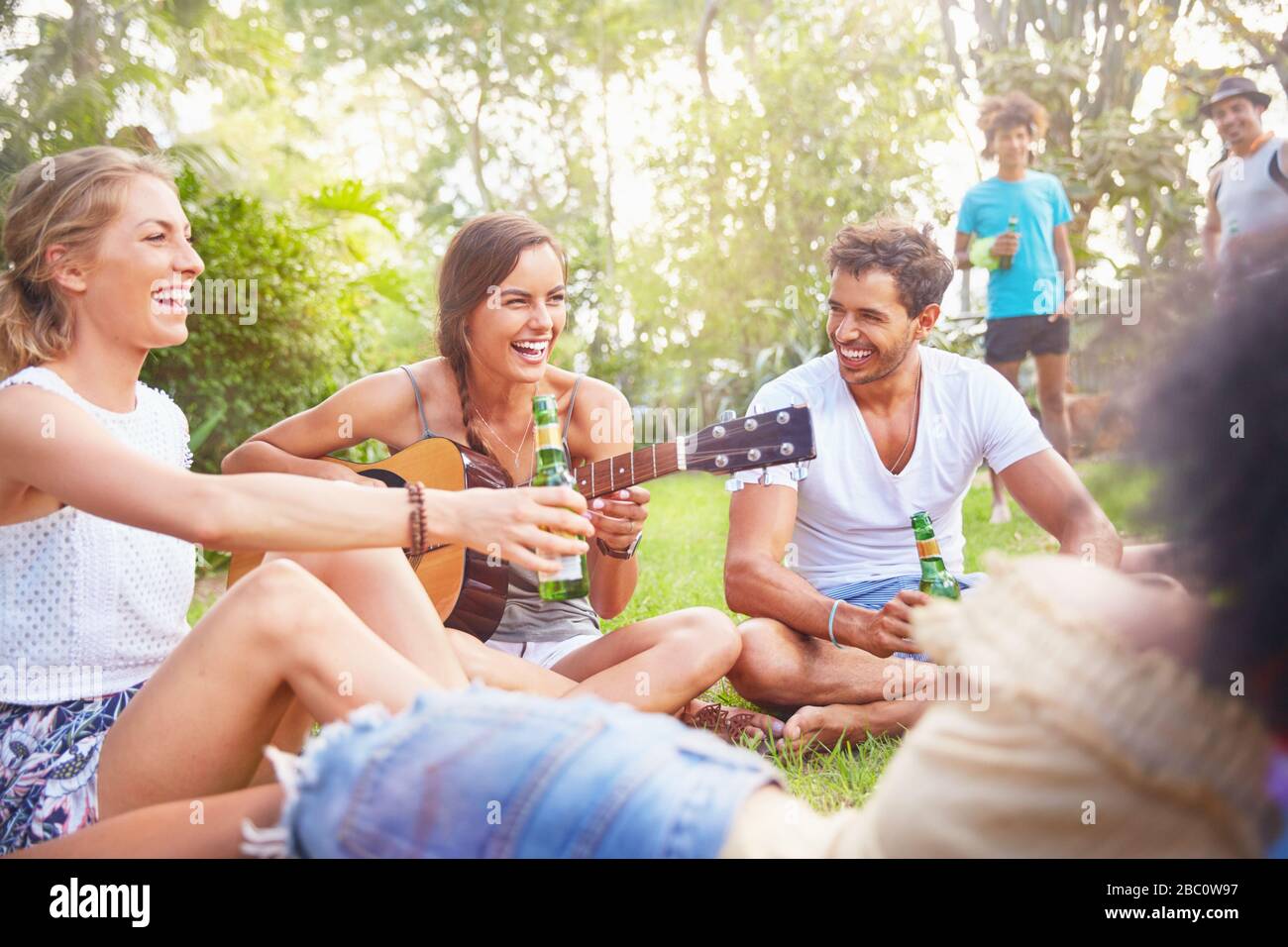 Young friends laughing, hanging out drinking and playing guitar in summer park Stock Photo