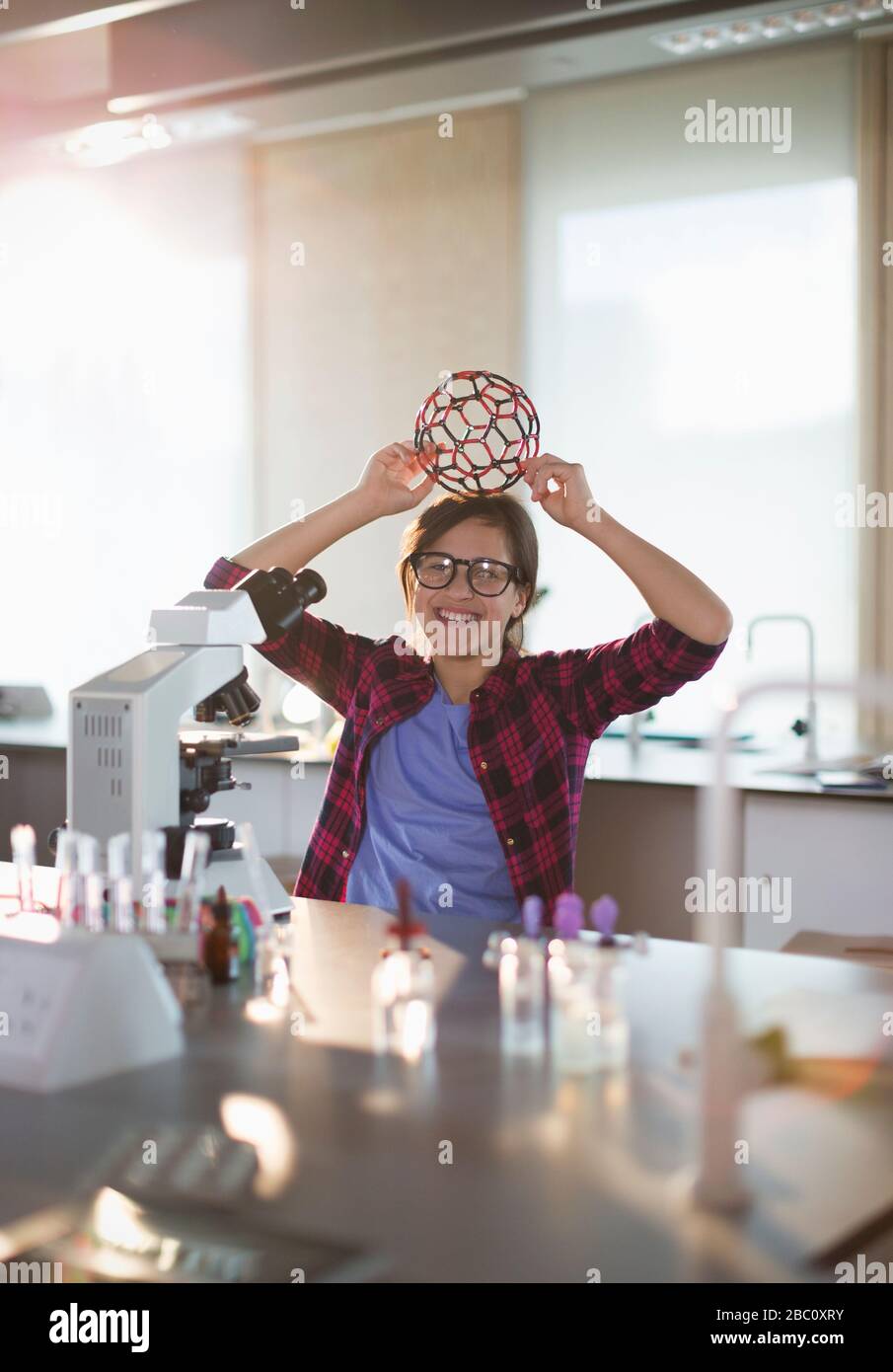 Portrait playful girl student holding molecular structure on head at microscope in laboratory classroom Stock Photo