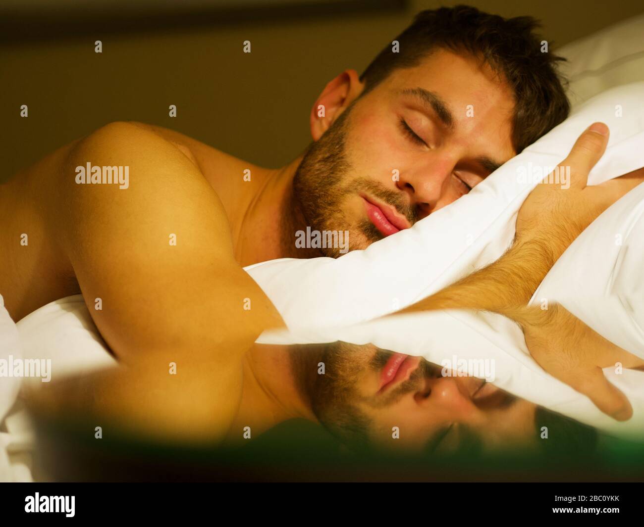 Closeup of a man sleeping in a bed with white sheets and reflection on the nightstand. Stock Photo