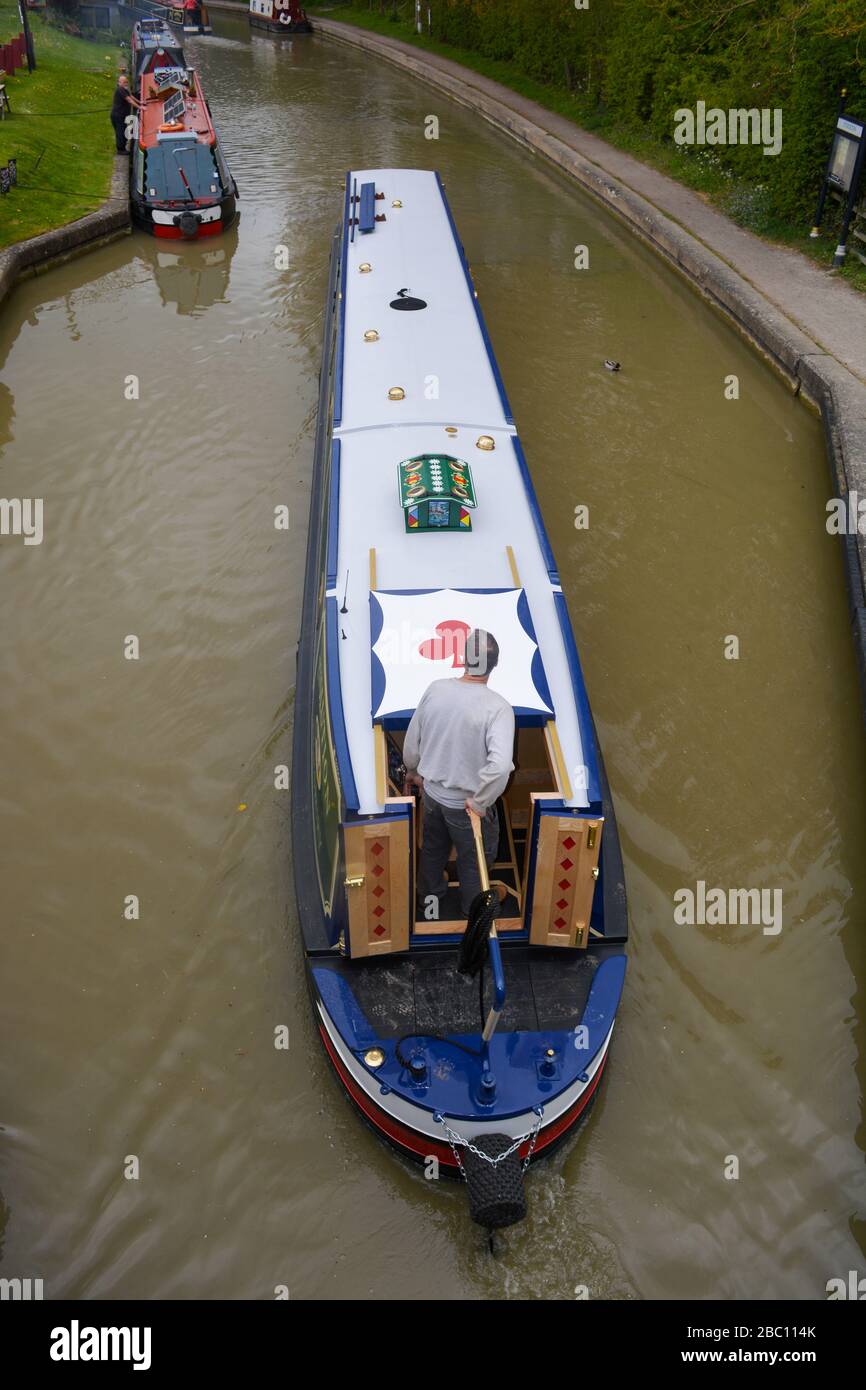 View from a road bridge looking down on top of a narrowboat passing beneath along the Grand Union Canal with a man at the tiller. Stock Photo