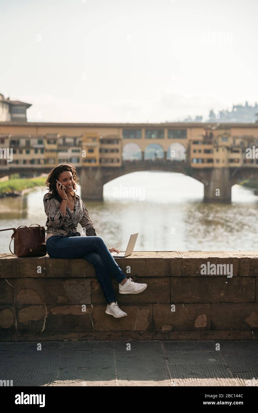 Young woman using laptop and cell phone on a bridge, Florence, Italy ...